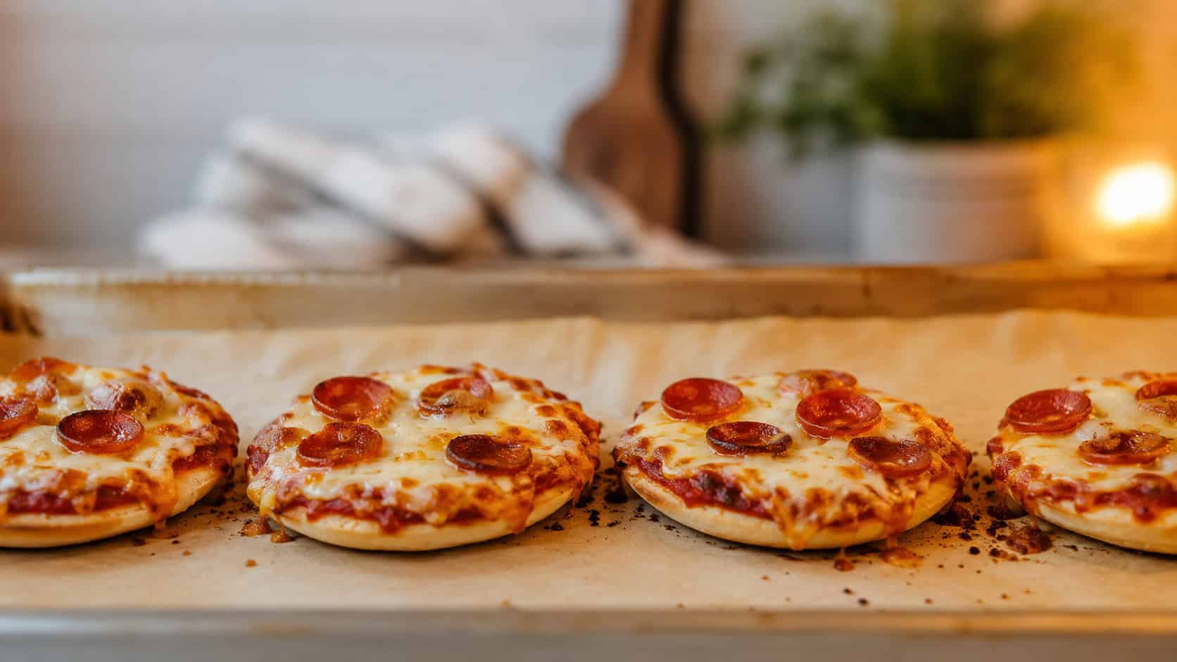 mini pita pizzas with bubbly melted cheese lined up in a wide horizontal row on a parchment-lined sheet pan, golden edges and bits of pepperoni, cozy kitchen light, shallow depth of field, landscape orientation