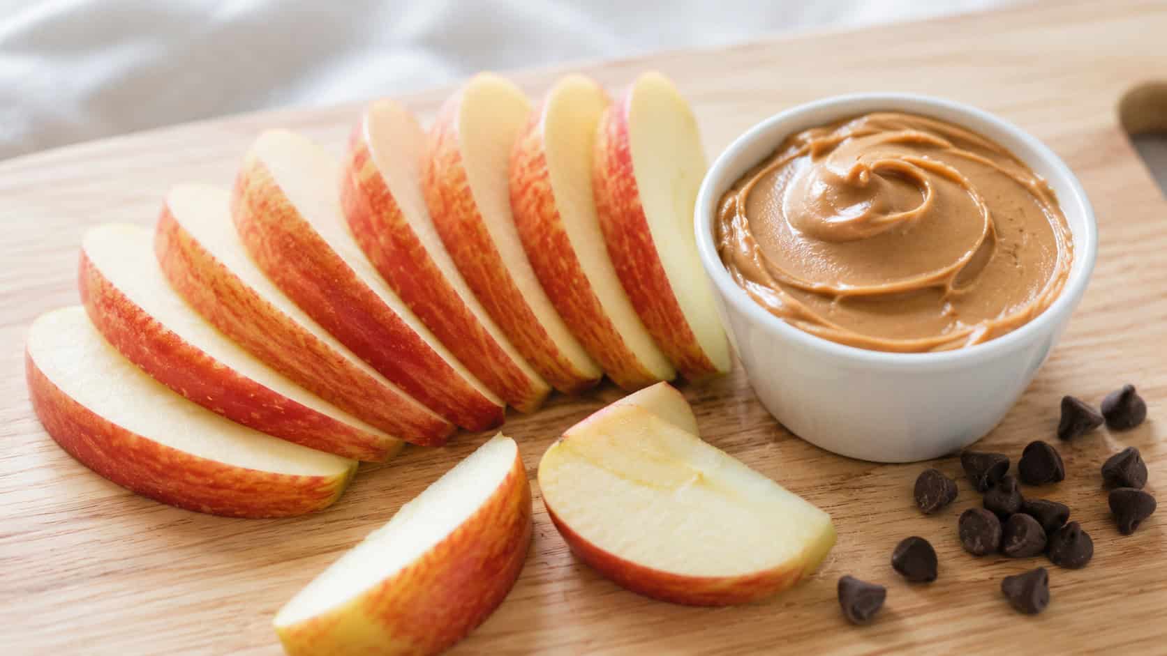 crisp apple slices fanned out in a wide horizontal arrangement beside a small cup of creamy peanut butter on a light wooden board, a few loose slices and a scatter of chocolate chips, bright natural light, shallow depth of field, landscape orientation