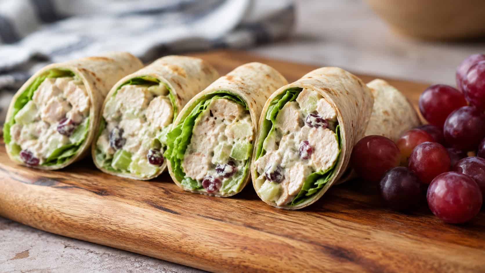 chicken salad wraps sliced and arranged in a wide horizontal row on a wooden board, creamy filling visible at the cut ends, a handful of grapes beside them, bright natural light, shallow depth of field, landscape orientation