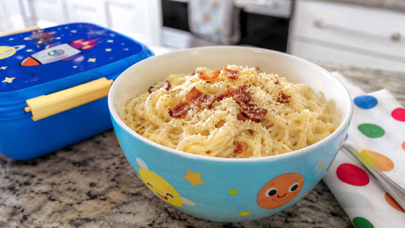 creamy cheesy ramen carbonara twirled in a kid-friendly bowl with crispy bacon bits and grated cheese on top, in a wide horizontal shot, a bright lunchbox and napkin beside it on a kitchen counter, natural light, shallow depth of field, landscape orientation