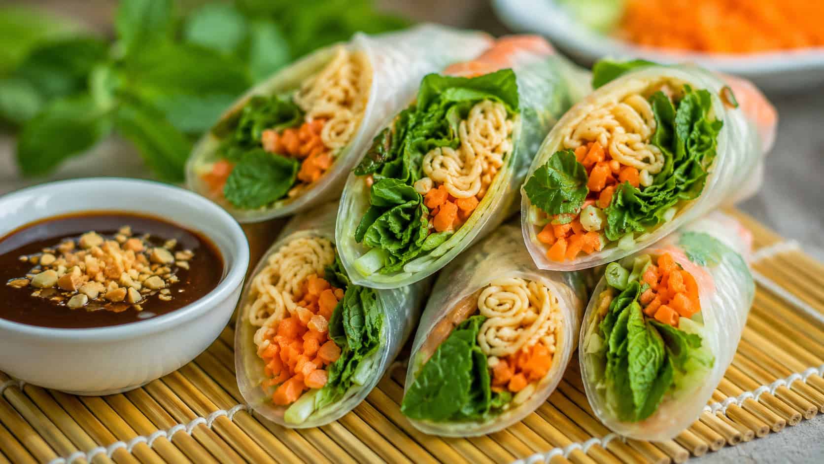 fresh ramen spring rolls cut on a diagonal to show colorful noodles, carrots, and mint inside translucent wrappers, in a wide horizontal shot, a small bowl of hoisin dipping sauce on a bamboo mat, bright natural light, shallow depth of field, landscape orientation