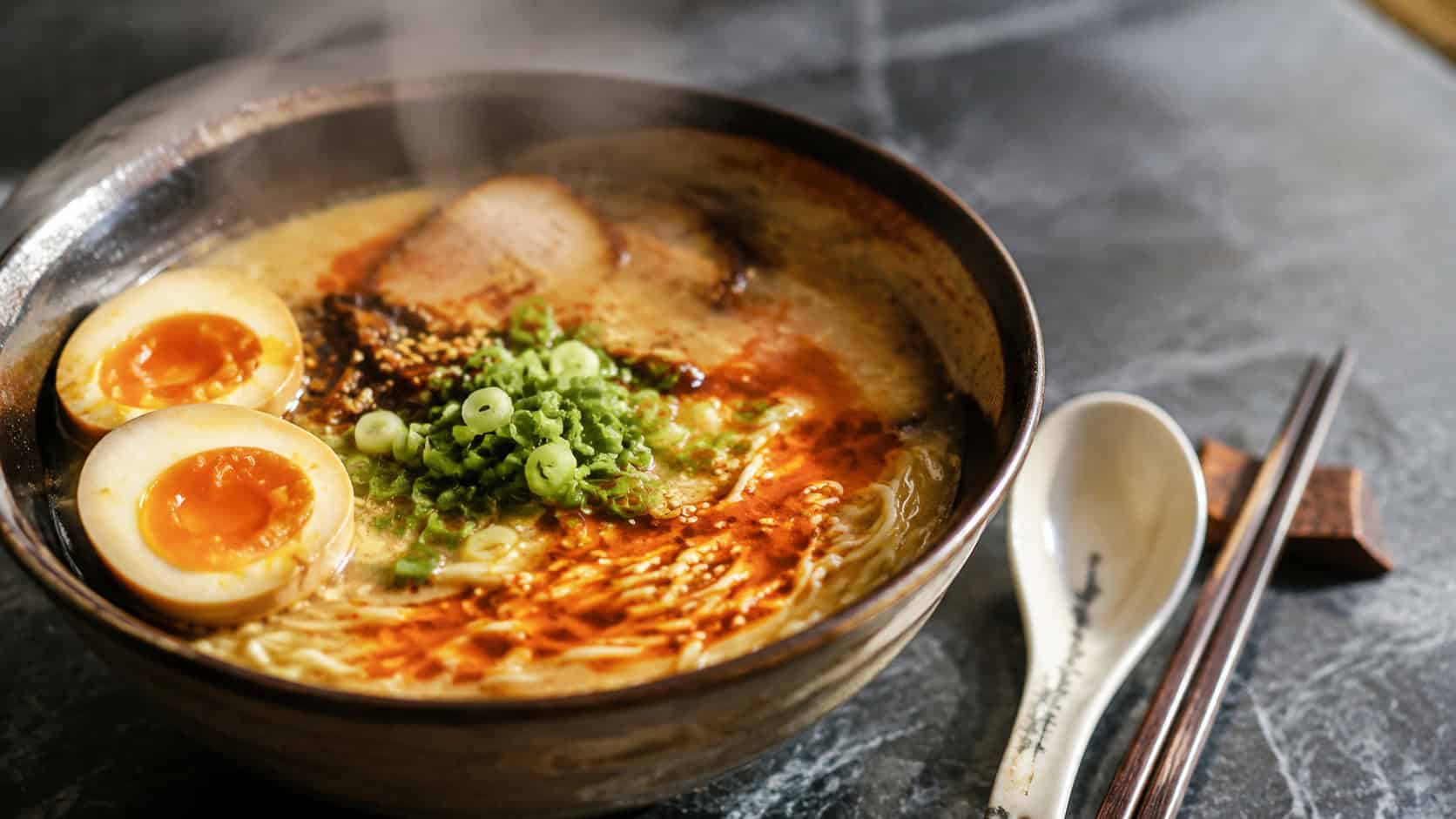 a steamy bowl of ramen with a halved jammy egg, green onions, and a swirl of chili oil, in a wide horizontal shot, a soup spoon and chopsticks beside it on a slate surface, warm natural light, shallow depth of field, landscape orientation