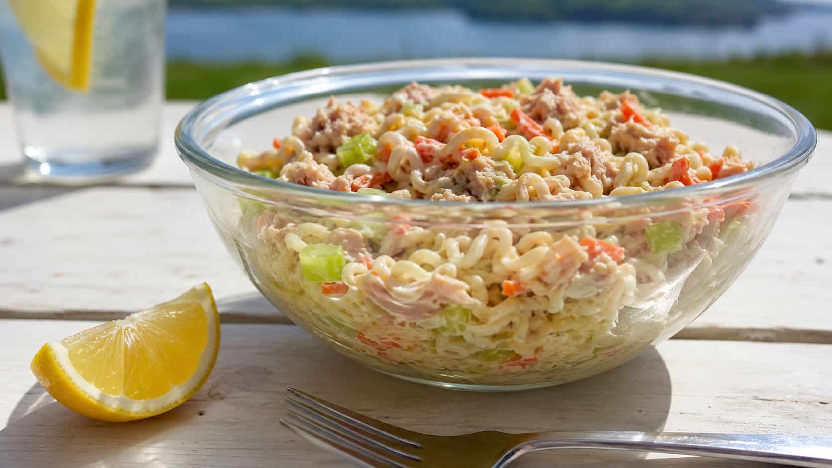 a chilled tuna ramen noodle salad with flecks of orange carrot and green celery in a clear bowl, in a wide horizontal shot, a lemon wedge and fork on a bright picnic table, fresh natural light, shallow depth of field, landscape orientation