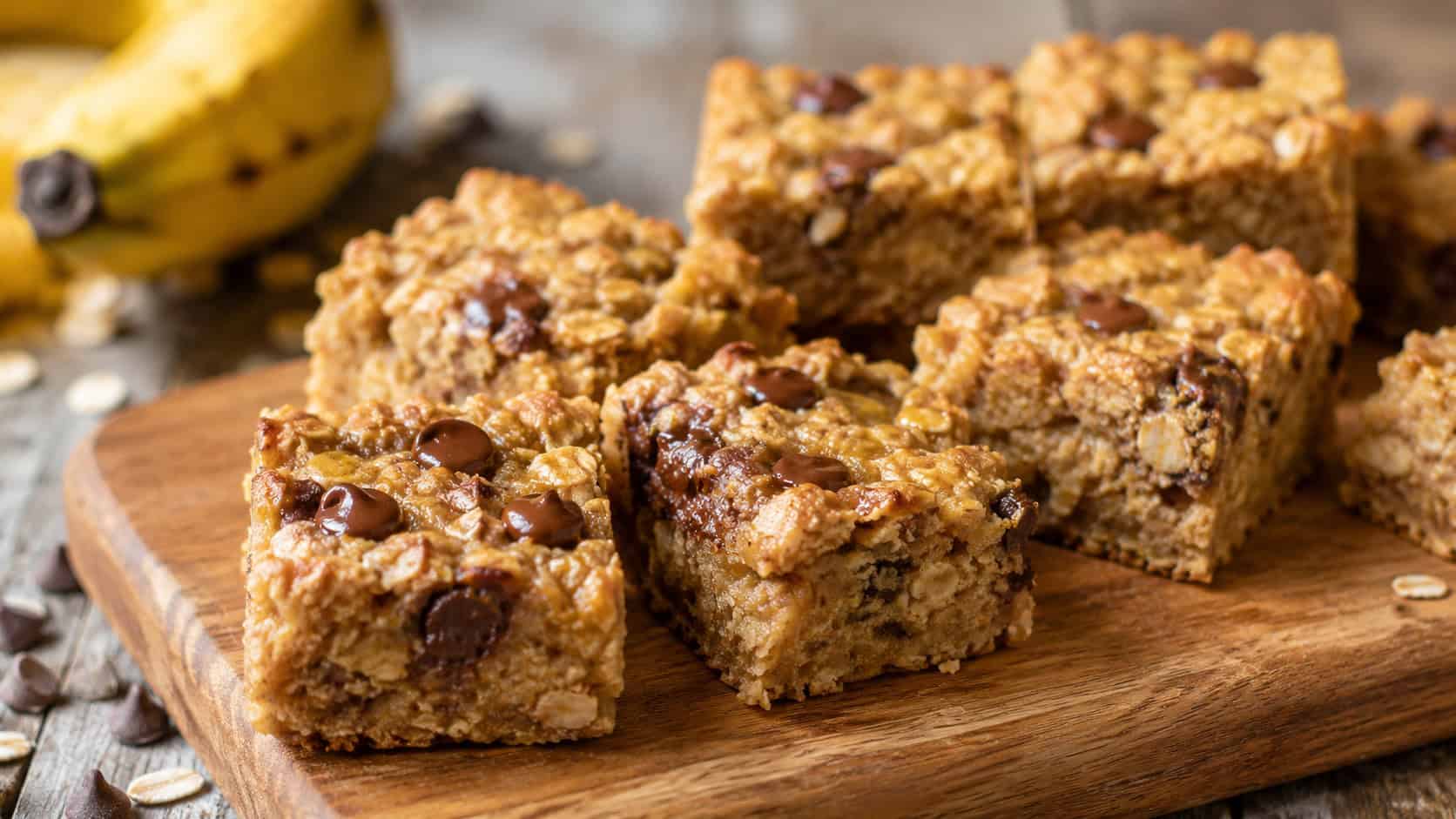 golden peanut butter banana oat bars cut into squares on a wooden board, a few studded with melty chocolate chips, in a wide horizontal shot on a rustic table, natural light, shallow depth of field, landscape orientation