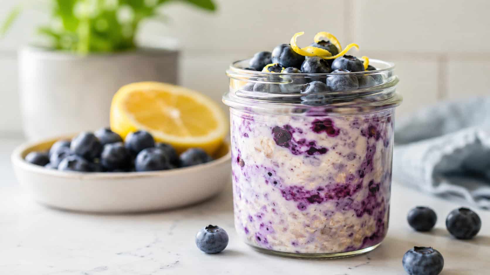 a jar of creamy blueberry overnight oats swirled purple and topped with whole blueberries and a curl of lemon zest, in a wide horizontal shot on a bright counter, natural light, shallow depth of field, landscape orientation