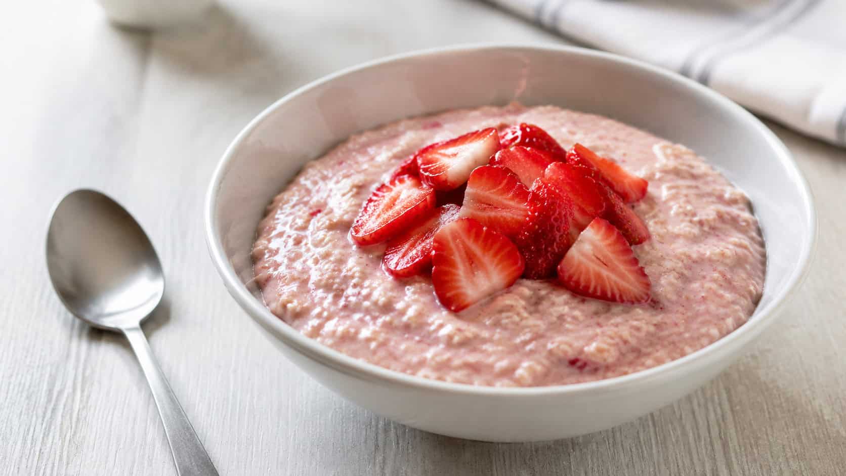 a creamy bowl of strawberry oatmeal tinged pink and topped with glossy sliced strawberries, in a wide horizontal shot on a light table, a spoon resting beside it, natural light, shallow depth of field, landscape orientation