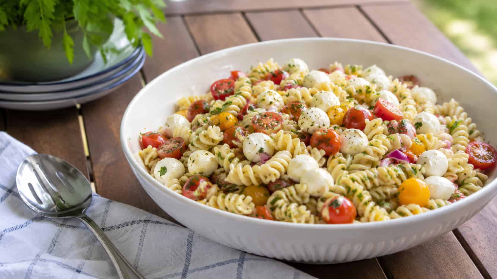 a soft pasta salad with rotini, mozzarella pearls, and tomatoes in a bowl, shown in a wide horizontal shot on a picnic table with a serving spoon nearby, natural light, shallow depth of field, landscape orientation
