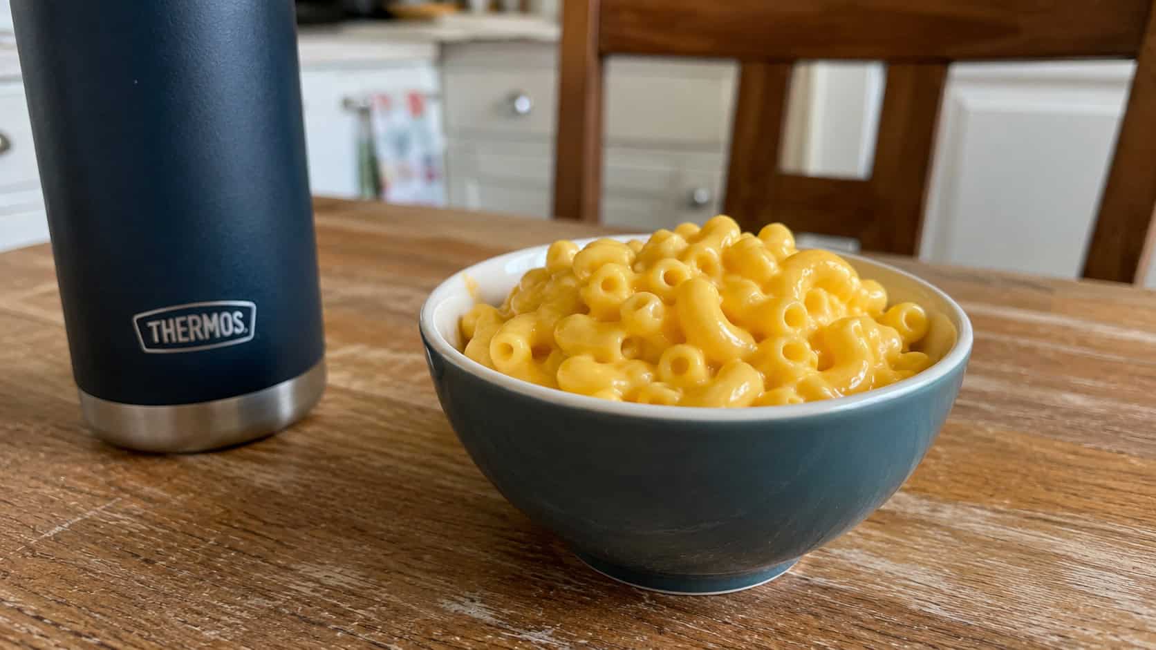 creamy macaroni and cheese with a glossy cheese sauce in a small bowl, shown in a wide horizontal shot on a kitchen table with a thermos beside it, natural light, shallow depth of field, landscape orientation