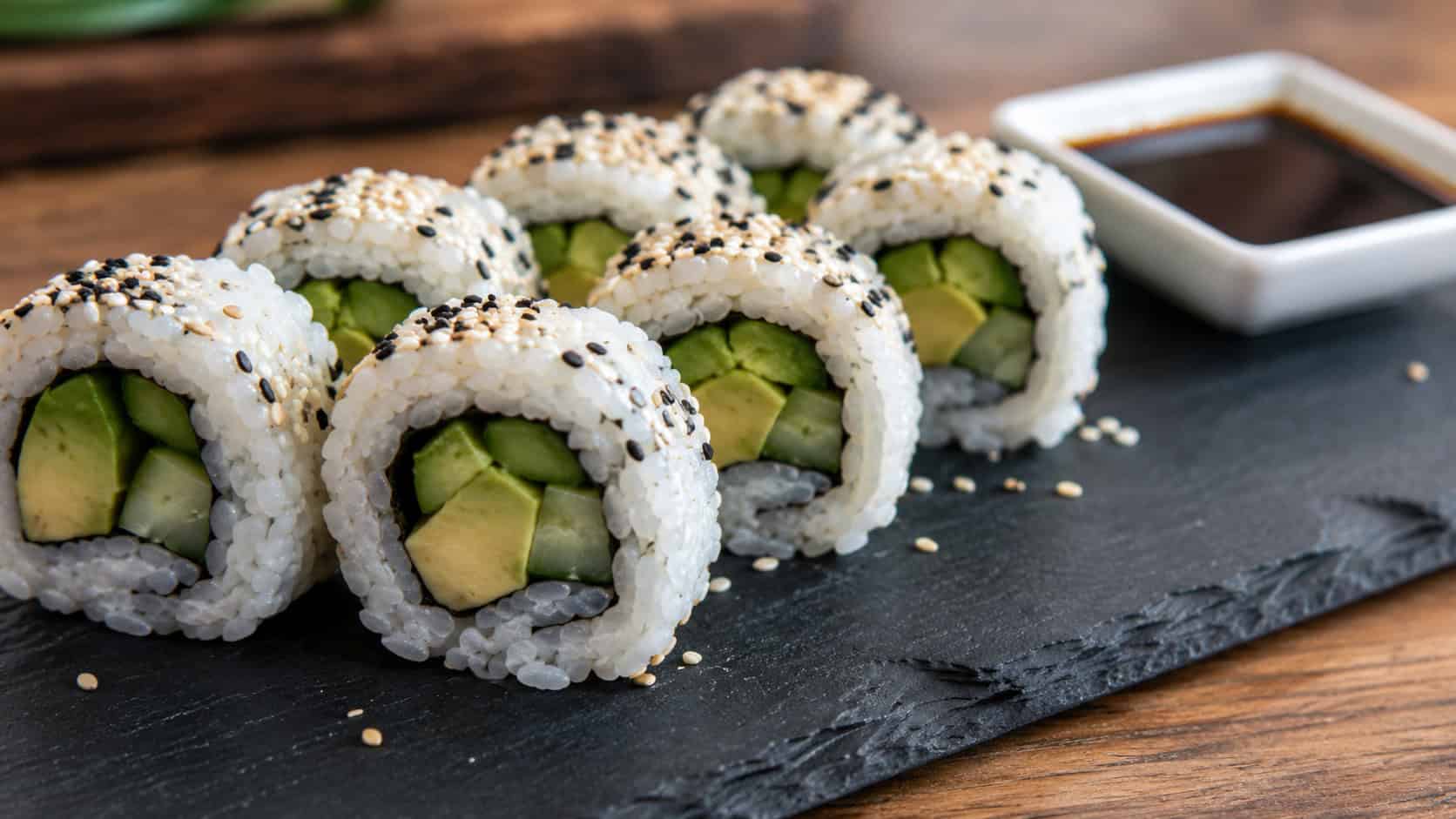 veggie sushi rolls cut into rounds showing avocado and cucumber centers, shown in a wide horizontal shot on a slate board with a small dish of soy sauce nearby, natural light, shallow depth of field, landscape orientation