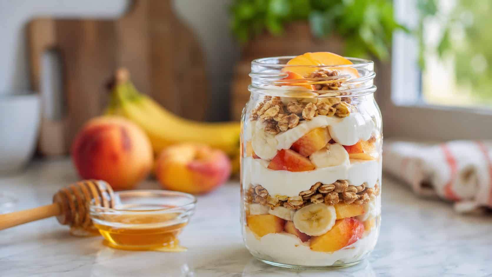 a layered yogurt parfait with soft banana and peach in a clear jar, shown in a wide horizontal shot on a bright counter with a drizzle of honey nearby, natural light, shallow depth of field, landscape orientation