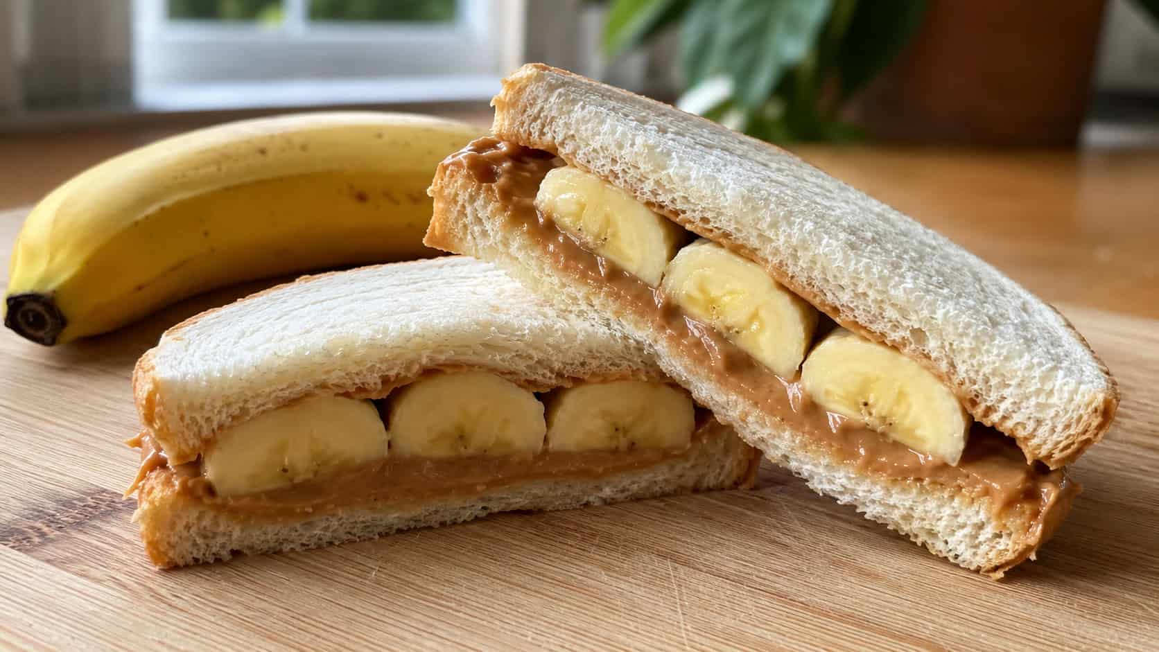 a soft peanut butter and banana sandwich cut in half to show the banana slices inside, shown in a wide horizontal shot on a wooden board with a whole banana nearby, natural light, shallow depth of field, landscape orientation