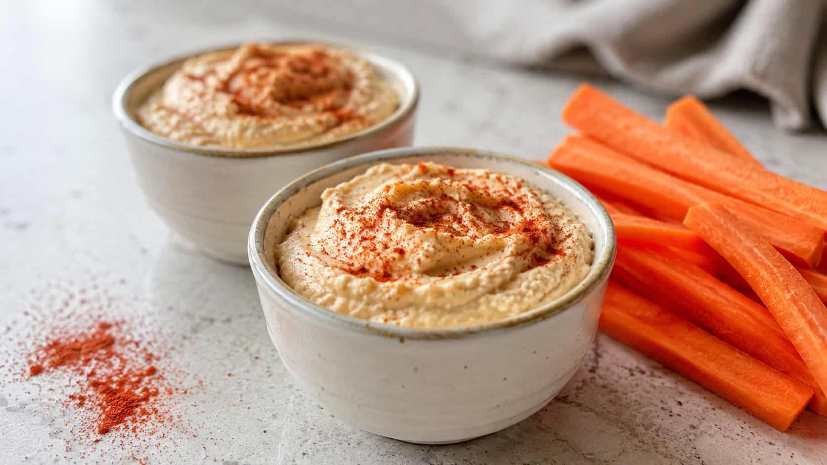 small cups of creamy hummus dusted with paprika beside soft carrot sticks, shown in a wide horizontal shot on a light counter with a sprinkle of paprika nearby, natural light, shallow depth of field, landscape orientation
