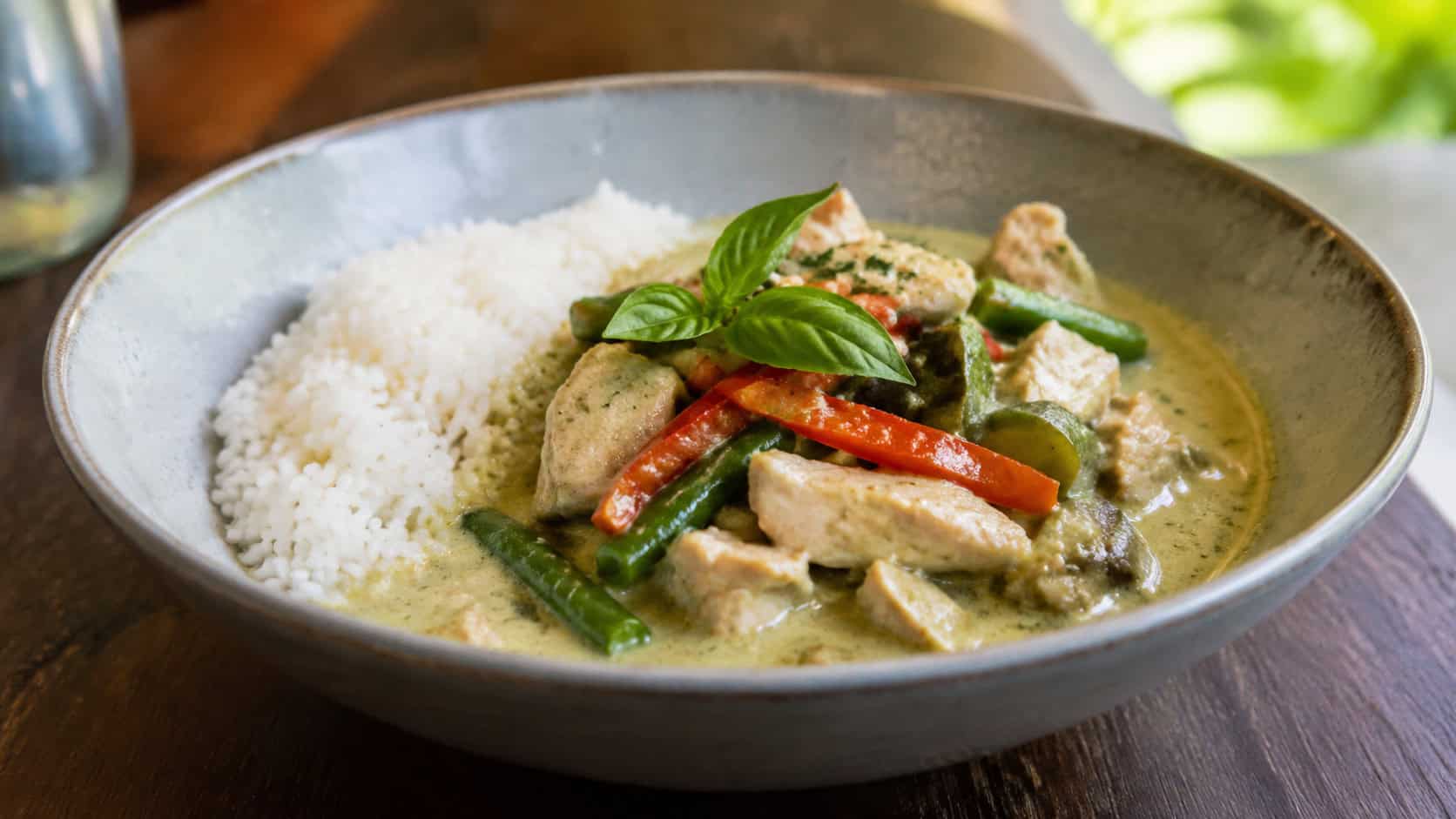 Thai green curry over rice in a wide horizontal shot, creamy pale-green coconut curry with bell peppers, green beans and chicken, in a bowl on a wooden table, natural light, shallow depth of field, landscape orientation
