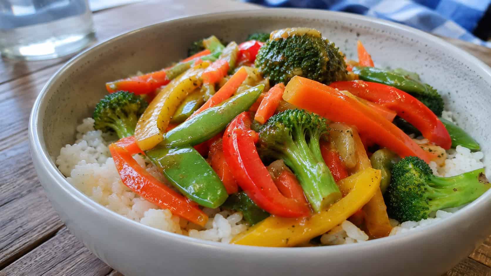a colorful vegetable stir fry over rice in a wide horizontal shot, glossy bell peppers, broccoli, snap peas and carrots, in a bowl on a wooden surface, natural daylight, shallow depth of field, landscape orientation