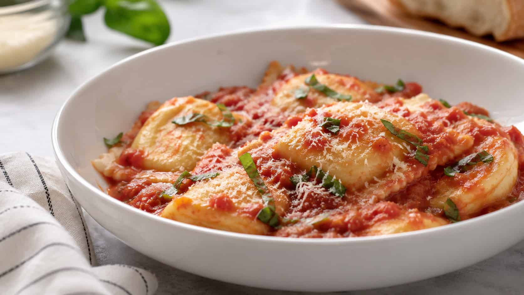 cheese ravioli in marinara sauce in a wide horizontal shot, plump pasta pillows coated in red sauce with grated parmesan and basil, in a bowl on a light table, natural light, shallow depth of field, landscape orientation