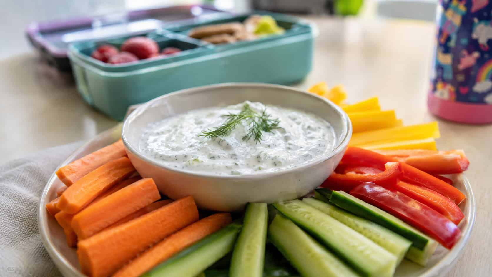 a bowl of creamy herb yogurt dip shot wide in landscape orientation on a bright table, surrounded by colorful carrot, cucumber, and pepper sticks, natural light, shallow depth of field, a lunchbox blurred in the background