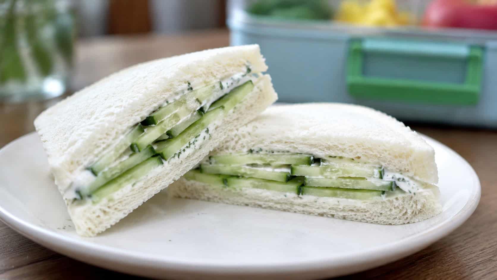 a yogurt and cucumber sandwich cut into halves shot wide in landscape orientation on a plate, soft bread with creamy spread and crisp cucumber layers visible, natural light, shallow depth of field, a lunchbox blurred in the background