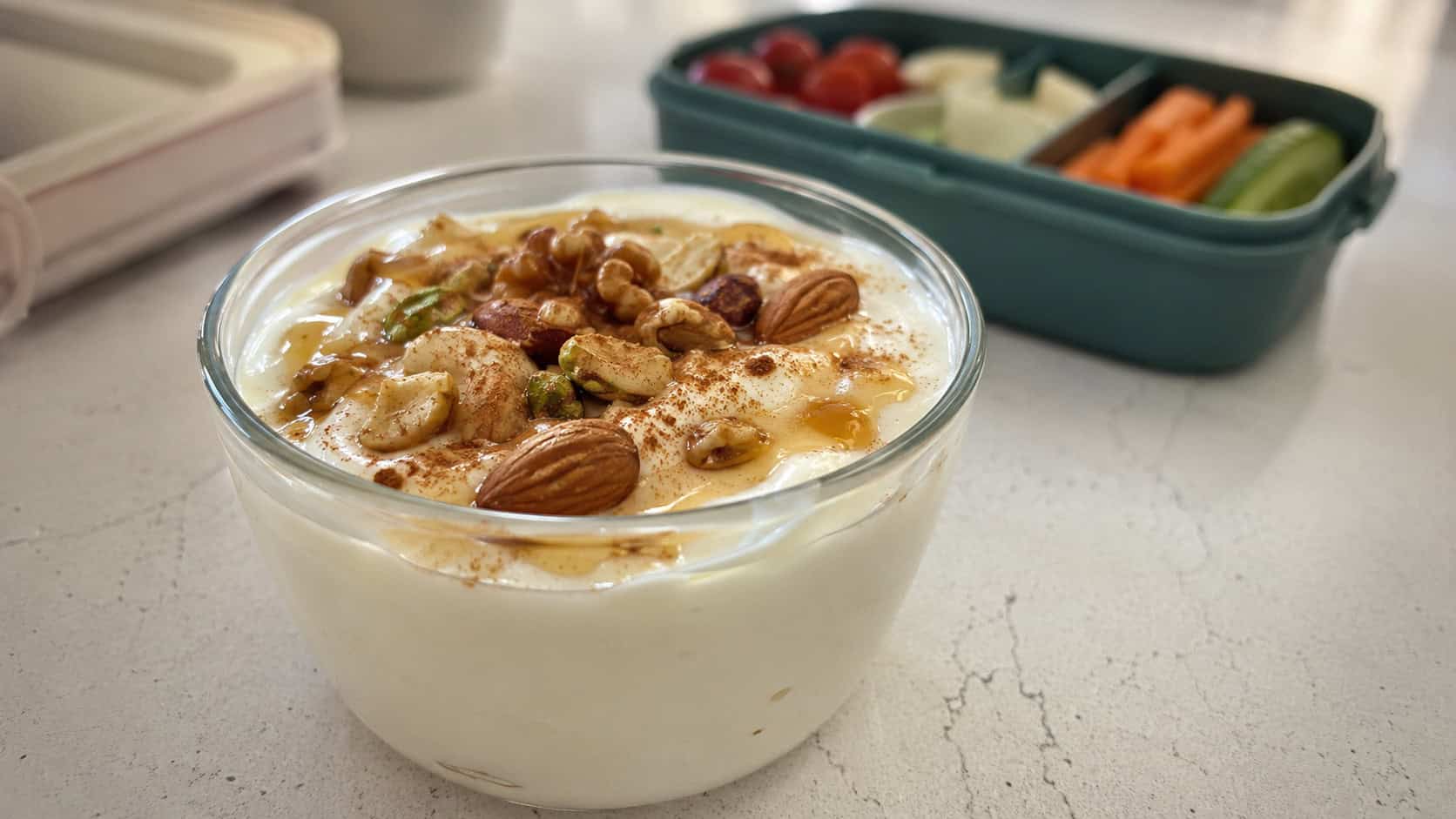 a cup of thick yogurt topped with mixed nuts and honey shot wide in landscape orientation on a bright counter, glossy honey drizzle and a dusting of cinnamon, natural light, shallow depth of field, a lunchbox blurred in the background