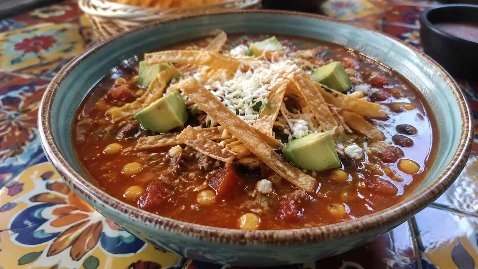 Mexican beef tortilla soup in a wide horizontal shot, red broth with ground beef and corn topped with crispy tortilla strips, avocado and cheese, in a bowl on a colorful table, natural light, shallow depth of field, landscape orientation