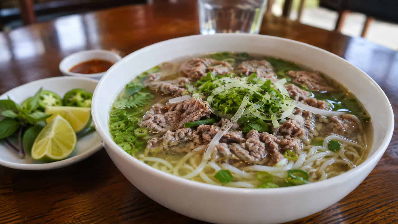 beef pho with noodles in a wide horizontal shot, clear aromatic broth with ground beef, rice noodles and fresh herbs, in a deep bowl with lime, on a wooden table, natural light, shallow depth of field, landscape orientation