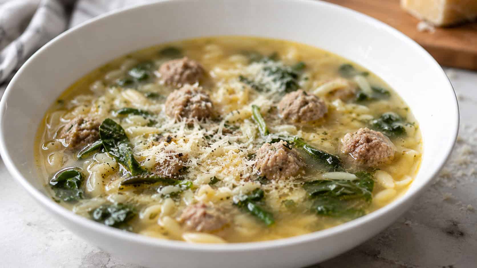 Italian wedding soup in a wide horizontal shot, golden broth with tiny meatballs, small pasta and green spinach, in a white bowl with parmesan, on a light table, natural light, shallow depth of field, landscape orientation