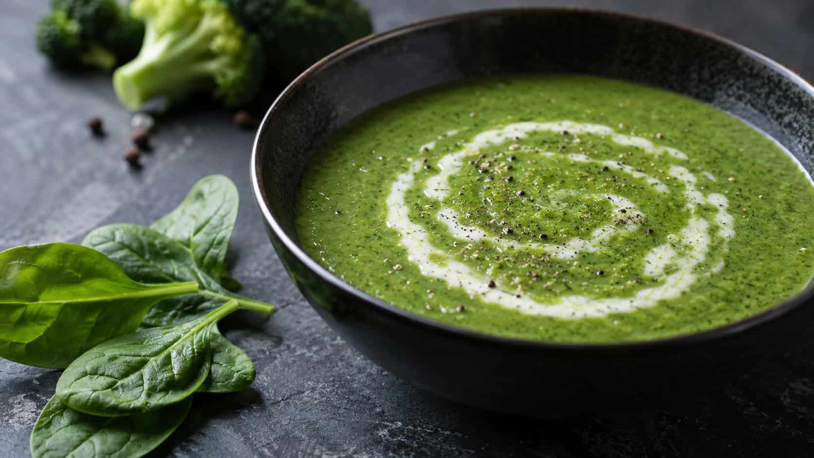 a deep-green broccoli and spinach soup in a dark bowl with a swirl of cream and pepper, in a wide horizontal shot, fresh spinach leaves beside it, on a moody slate table, natural light, shallow depth of field, landscape orientation