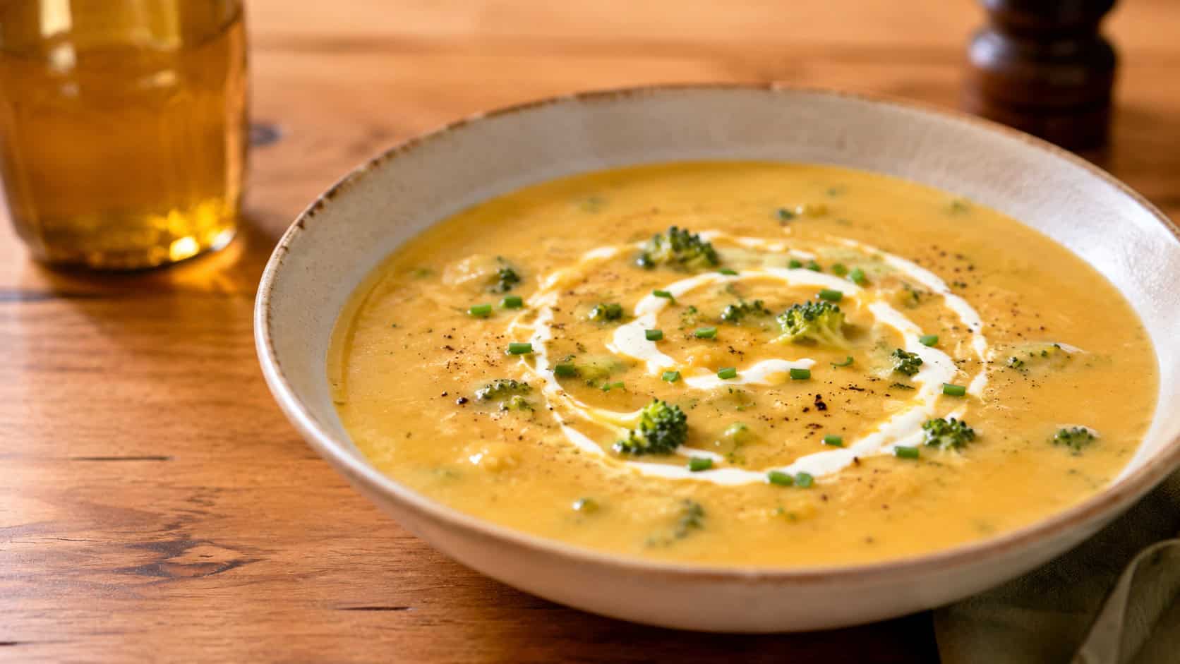 a velvety smooth broccoli cheddar bisque in a wide shallow bowl with a swirl of cream and chives, in a wide horizontal shot, golden and orange tones, on a warm wooden table, natural light, shallow depth of field, landscape orientation