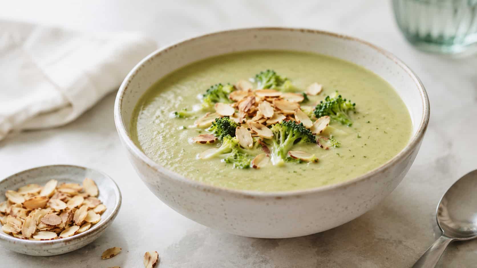 a creamy dairy-free broccoli soup in a light ceramic bowl topped with toasted slivered almonds, in a wide horizontal shot, a small dish of almonds nearby, on a bright minimalist table, natural light, shallow depth of field, landscape orientation