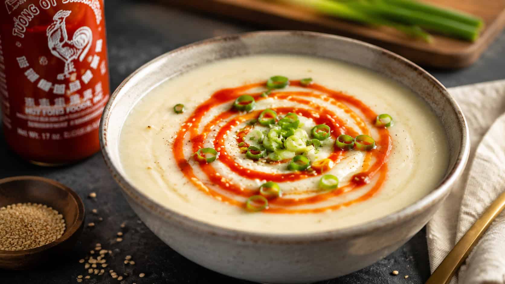 a creamy cauliflower soup with a fiery red sriracha swirl and green onion slices on top, in a wide horizontal shot, a bottle of chili sauce and sesame seeds on a dark table, warm natural light, shallow depth of field, landscape orientation