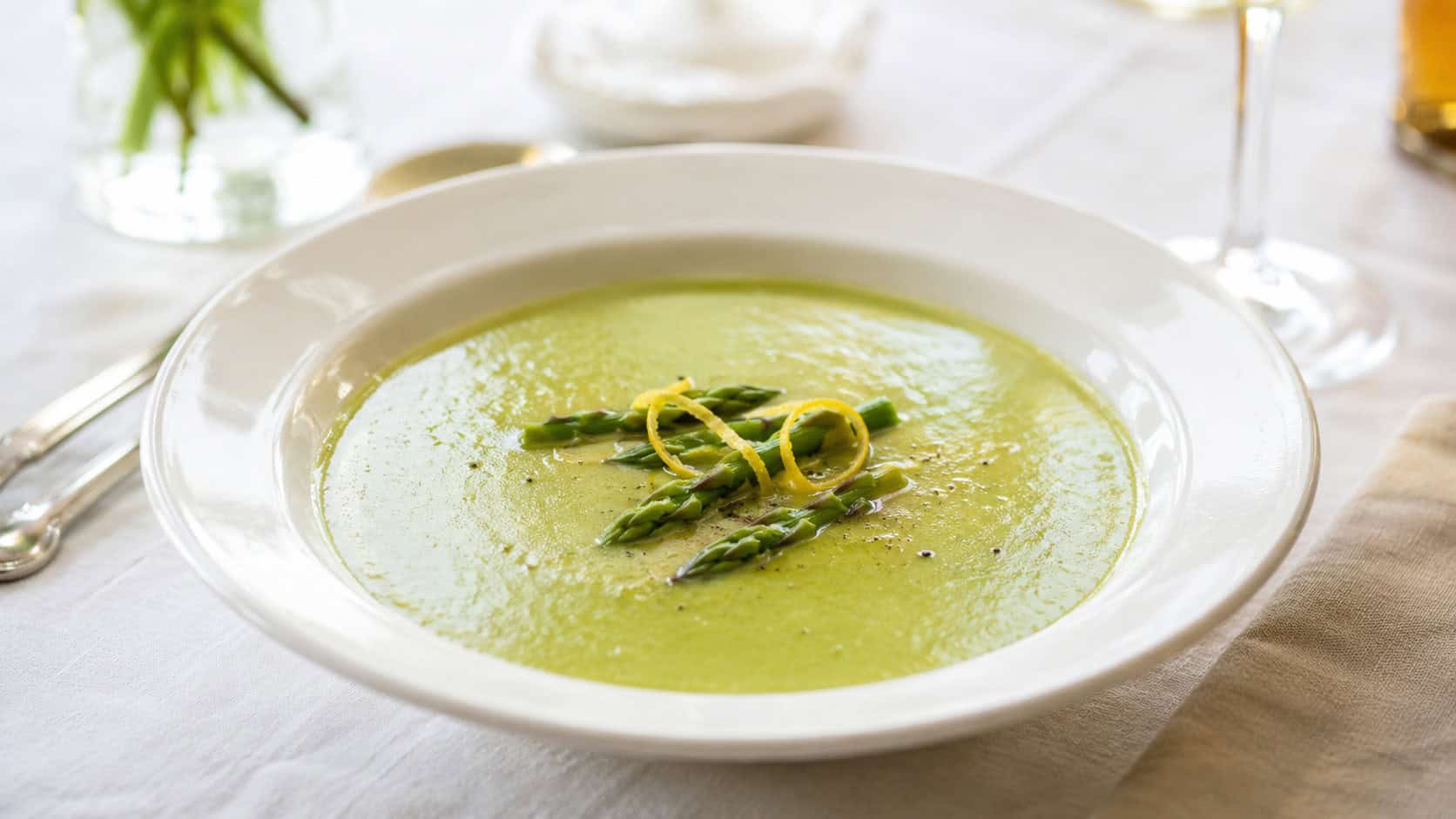 a bowl of smooth green asparagus soup in a wide horizontal shot, garnished with asparagus tips and a lemon twist, on a bright elegant table, fresh natural light, shallow depth of field, landscape orientation