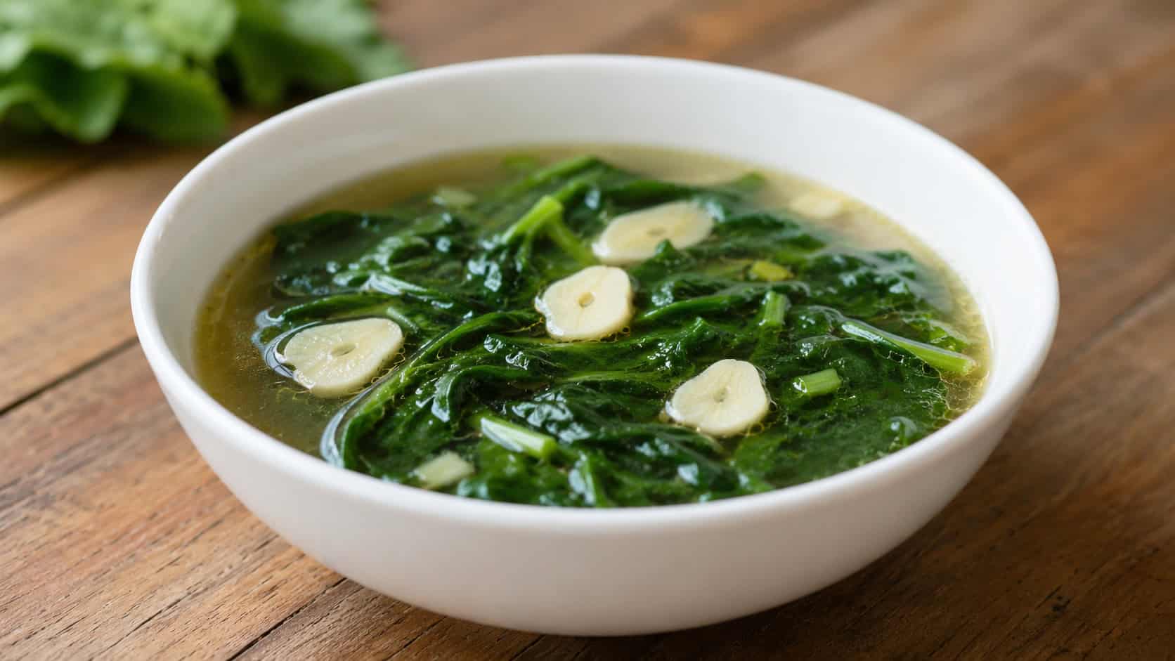 a bowl of clear garlic spinach broth in a wide horizontal shot, golden broth with wilted spinach and slices of garlic, in a simple white bowl on a wooden table, soft natural light, shallow depth of field, landscape orientation