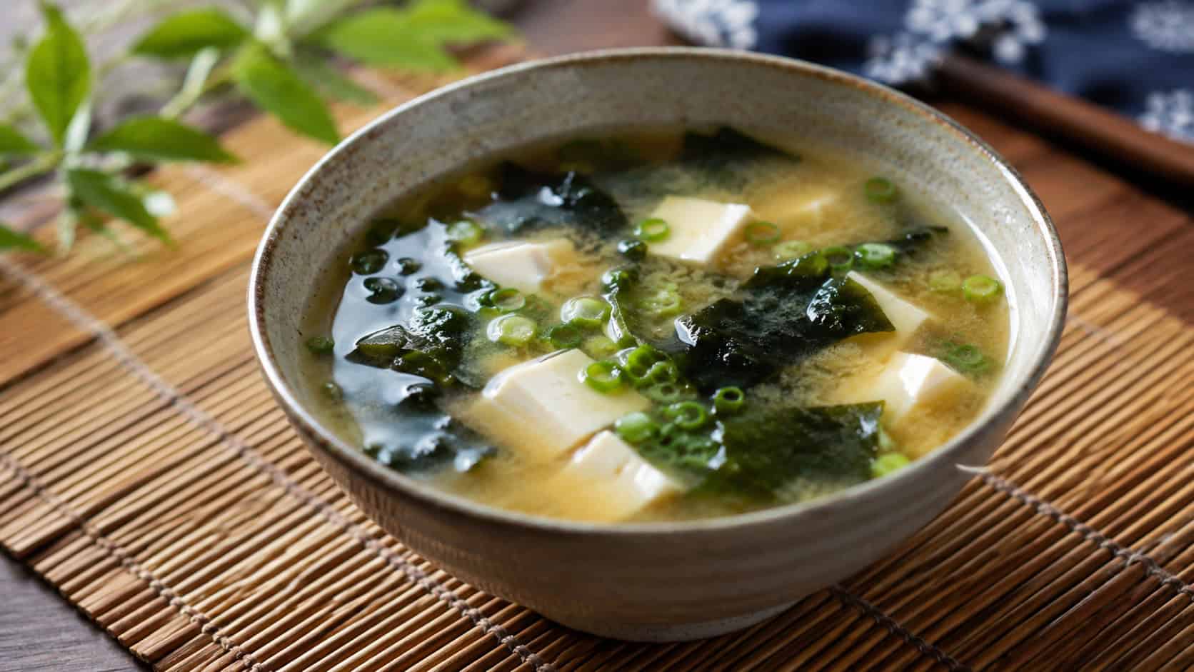 a bowl of miso soup in a wide horizontal shot, golden broth with cubes of tofu, dark seaweed, and green onion, in a ceramic bowl on a bamboo mat, soft natural light, shallow depth of field, landscape orientation