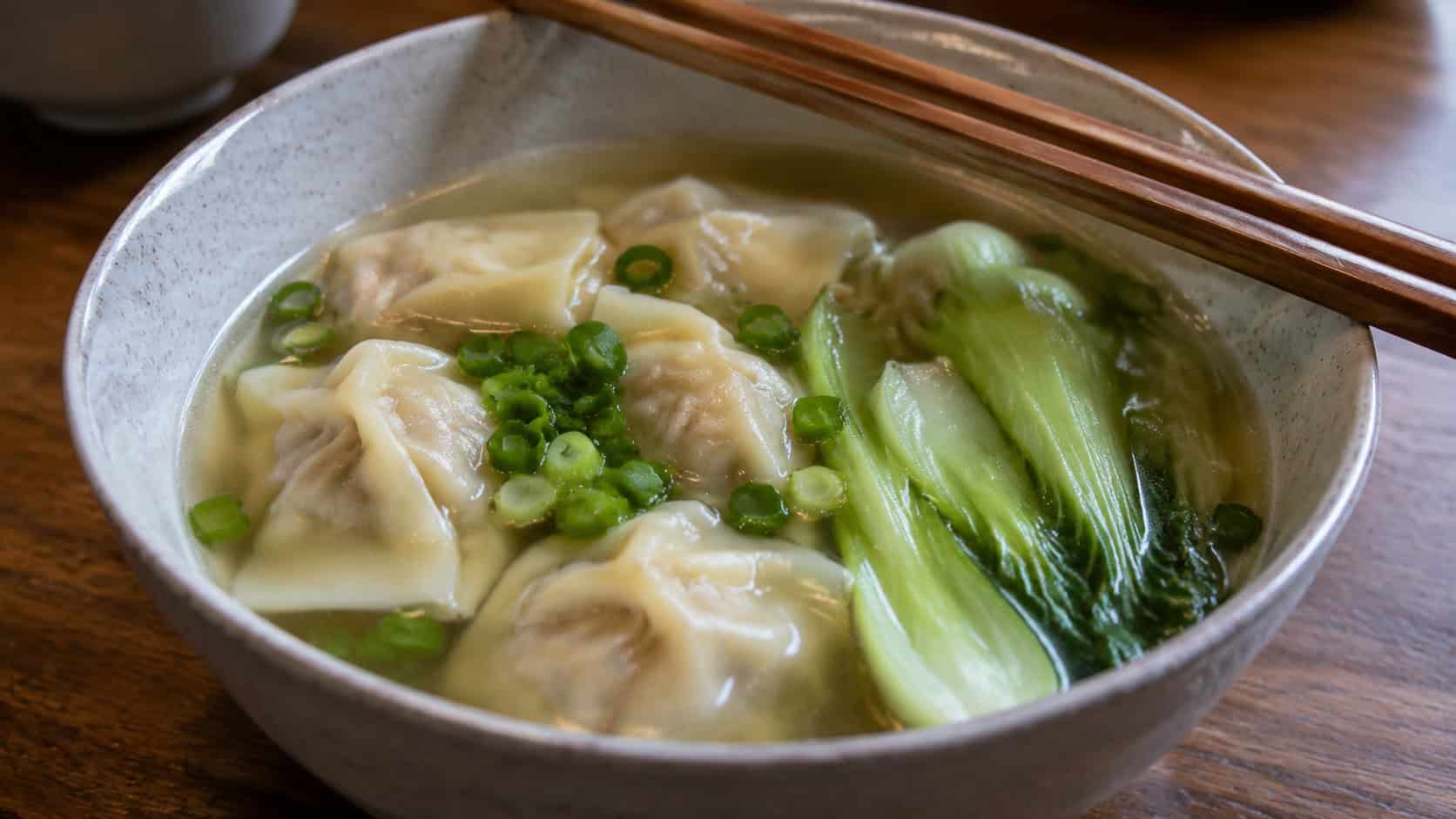 chicken wonton soup with plump wontons, halved bok choy, and green onions in a clear broth in a wide horizontal shot in a bowl, chopsticks resting on the rim, soft natural light, shallow depth of field, landscape orientation