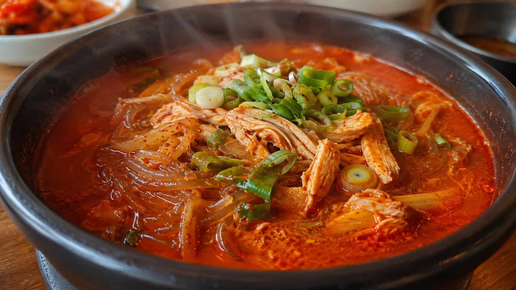 spicy Korean chicken soup with a fiery red broth, shredded chicken, glass noodles, and scallions in a wide horizontal shot in a bowl, steam rising, warm natural light, shallow depth of field, landscape orientation