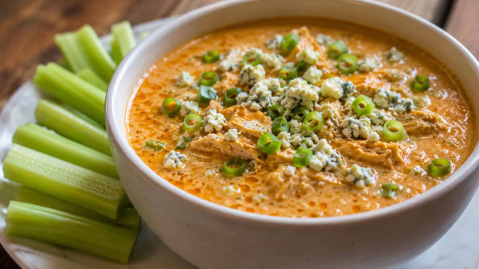 creamy orange buffalo chicken soup topped with blue cheese crumbles and green onions in a wide horizontal shot in a bowl, celery sticks on the side, warm natural light, shallow depth of field, landscape orientation