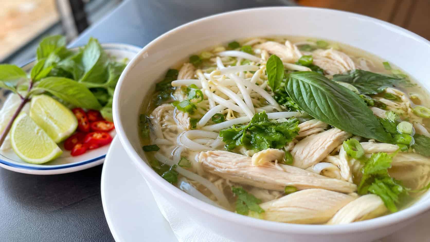 fragrant chicken pho with rice noodles, sliced chicken, bean sprouts, and Thai basil in a clear broth in a wide horizontal shot in a bowl, lime and chili on the side, natural light, shallow depth of field, landscape orientation