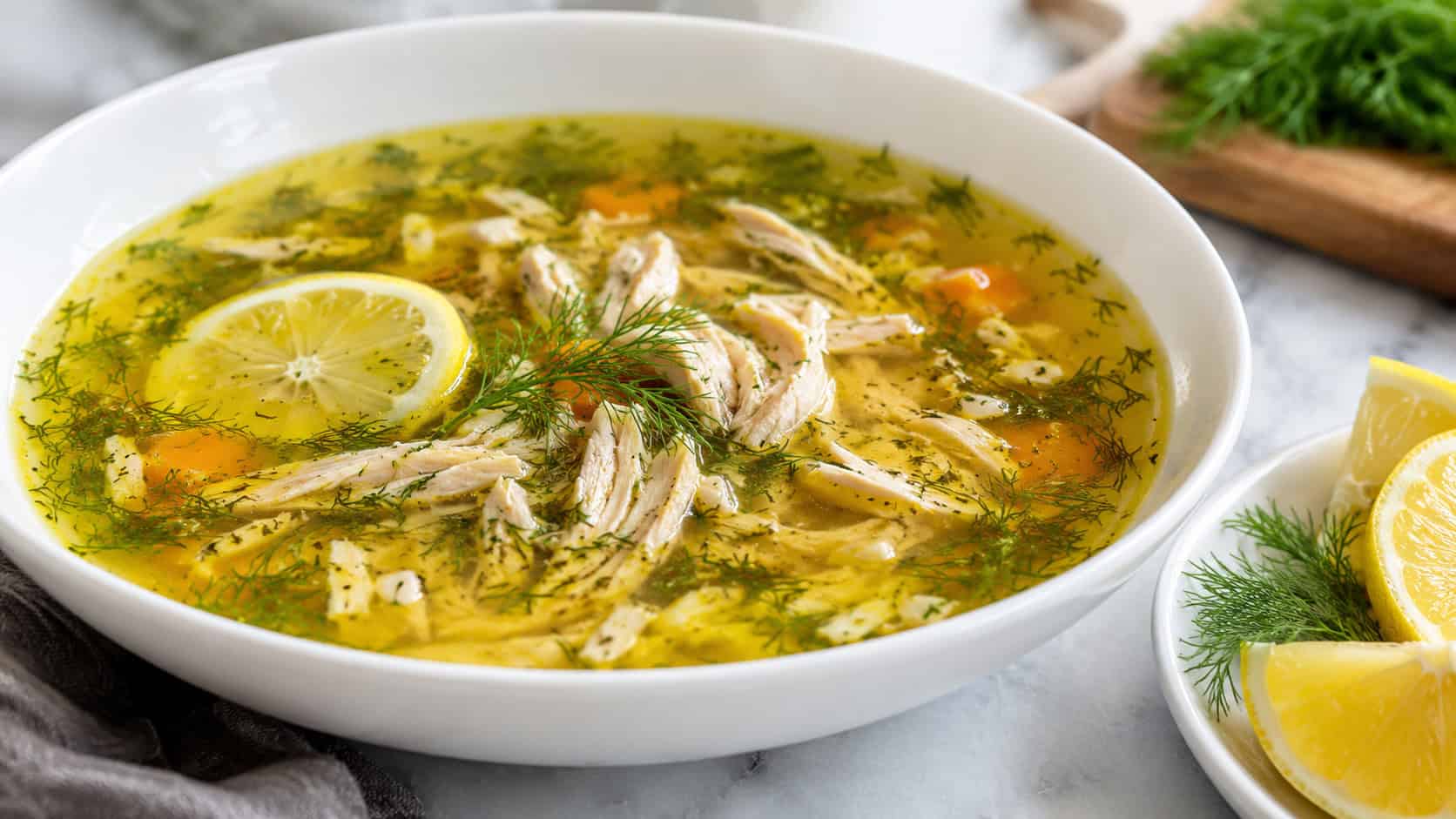 bright lemon herb chicken soup with a clear golden broth, shredded chicken, and fresh dill in a wide horizontal shot in a white bowl, lemon wedges beside it, natural light, shallow depth of field, landscape orientation
