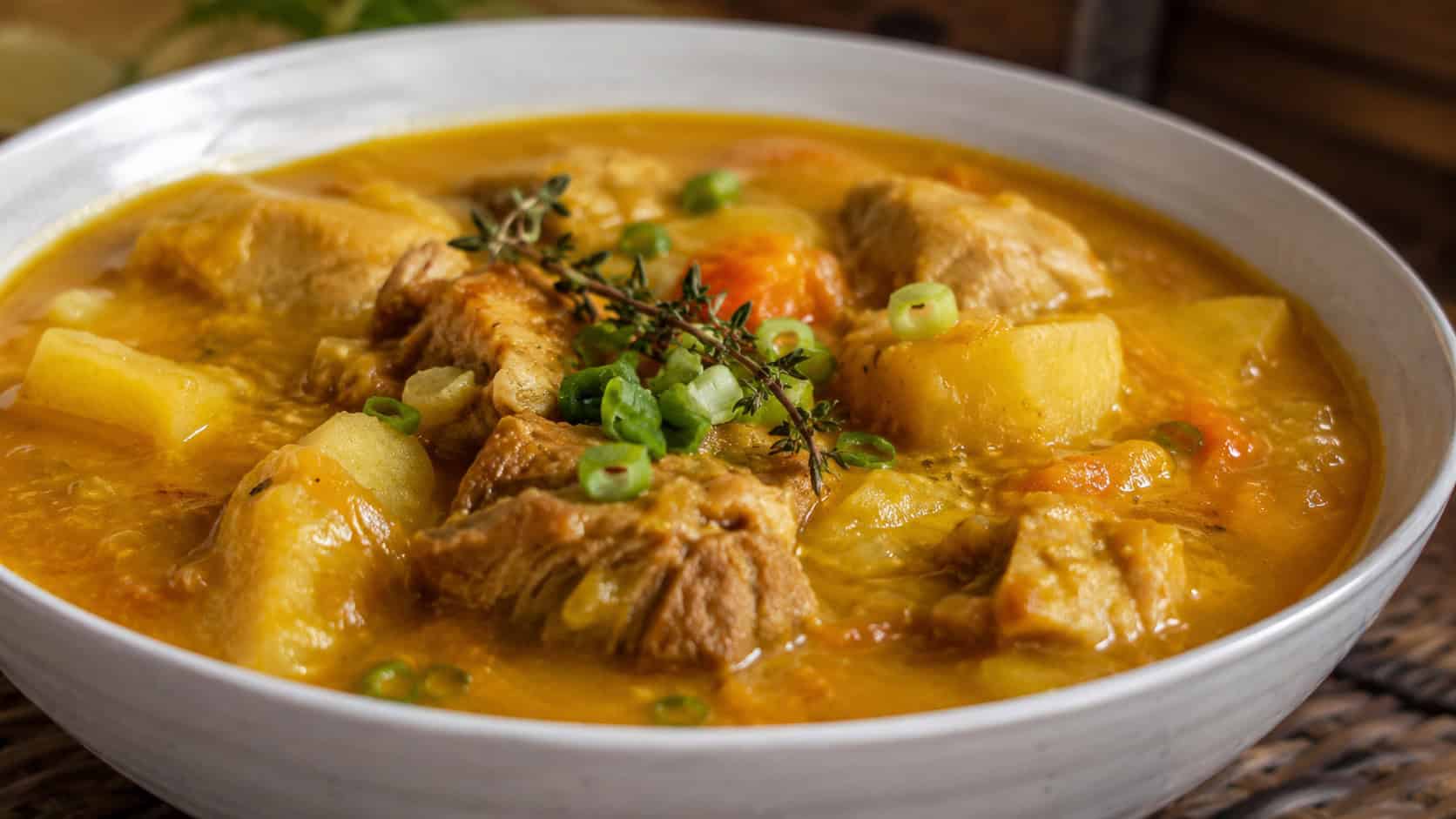 golden Jamaican chicken pumpkin soup with a thick orange broth, chicken, and potato in a wide horizontal shot in a bowl, fresh thyme and green onion on top, warm natural light, shallow depth of field, landscape orientation