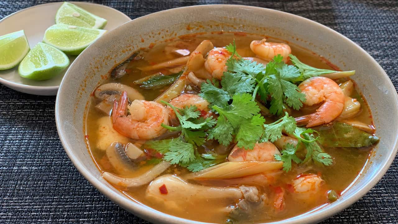 a tom yum soup in a wide bowl, clear reddish broth with pink shrimp, mushrooms, lemongrass, and fresh cilantro, lime wedges on the side, on a dark woven placemat, natural light, shallow depth of field, landscape orientation