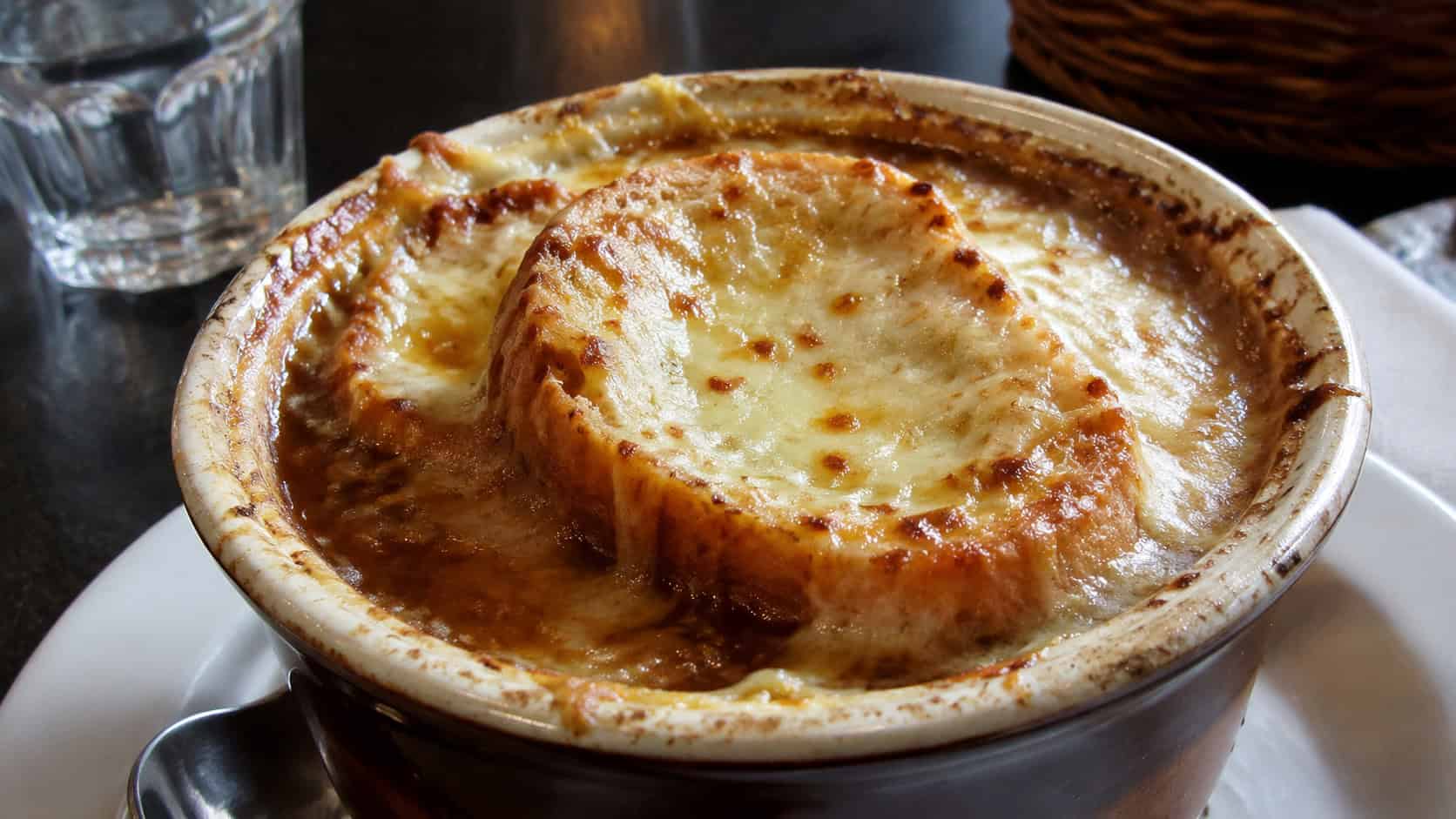 French onion soup in a wide crock, bubbling melted Gruyère over a golden toasted baguette slice, deep brown broth peeking through, on a dark bistro table, natural light, shallow depth of field, landscape orientation