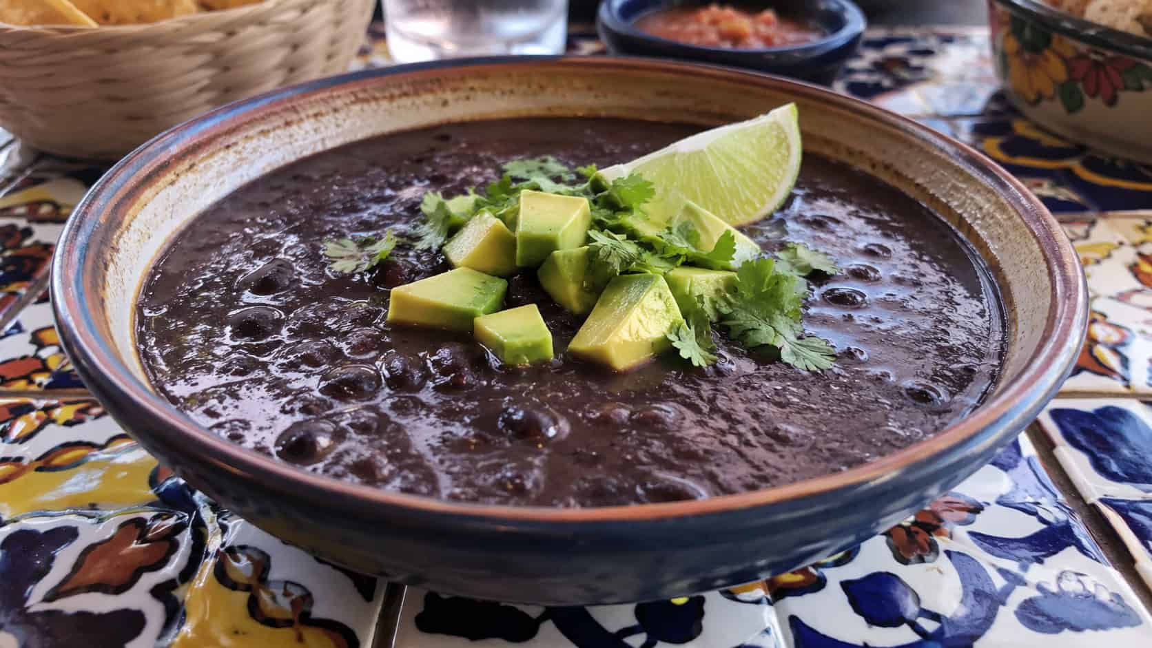 a thick smoky black bean soup in a wide bowl, dark and glossy, topped with diced avocado, cilantro, and a lime wedge, on a colorful tiled table, natural light, shallow depth of field, landscape orientation