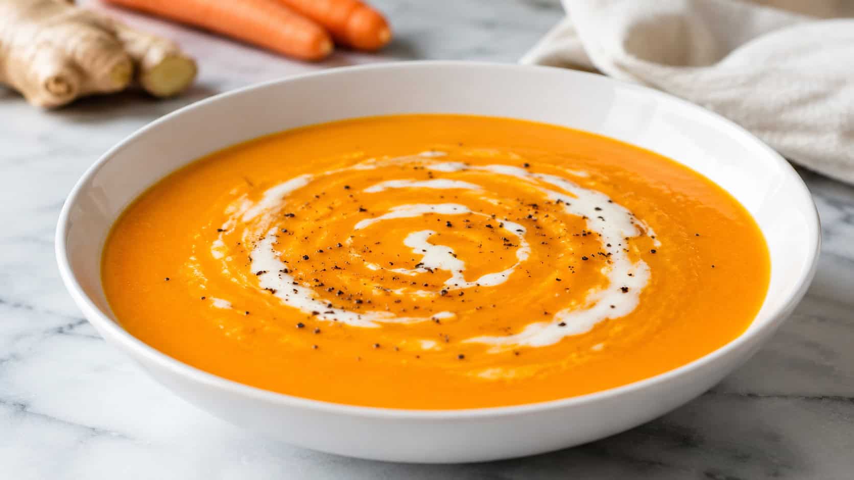 a smooth vibrant carrot ginger soup in a wide white bowl, bright orange with a swirl of coconut milk and a sprinkle of black pepper, on a light marble countertop, natural light, shallow depth of field, landscape orientation