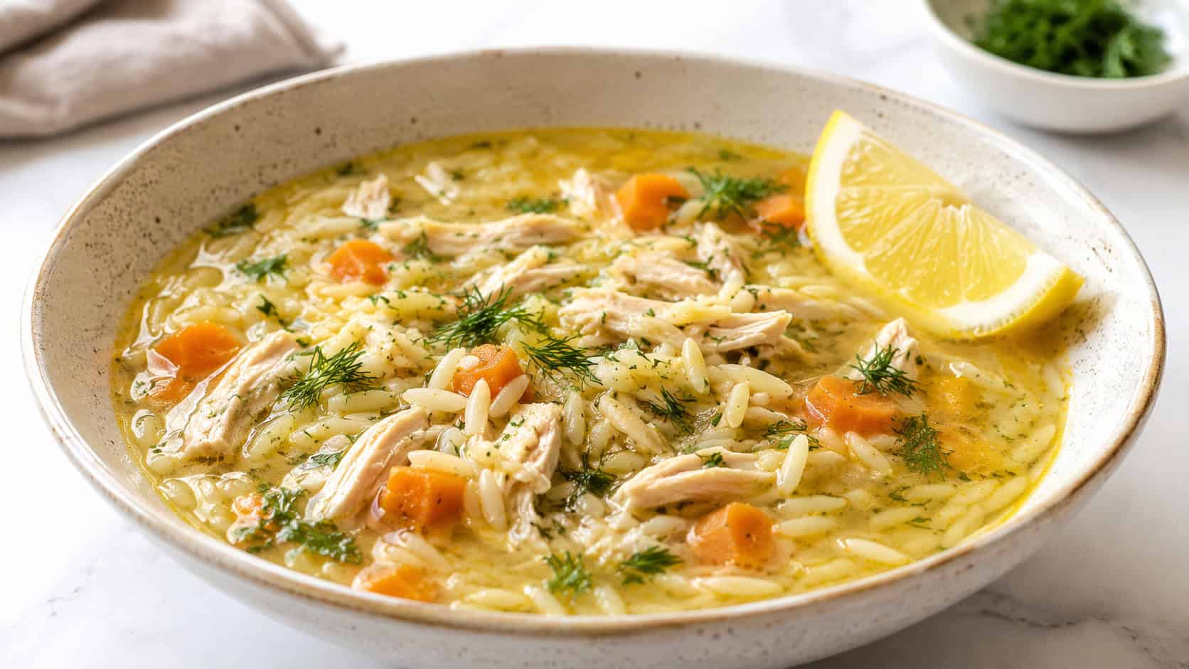 a lemony chicken orzo soup in a wide bowl, light golden broth with shredded chicken, orzo, carrots, and fresh dill, a lemon wedge on the side, on a bright white table, natural light, shallow depth of field, landscape orientation