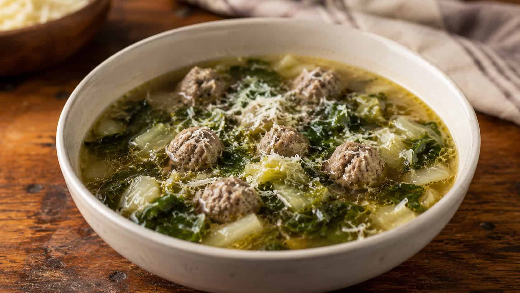 Italian meatball and escarole soup in a bowl, in a wide horizontal shot, small meatballs and leafy greens in a clear broth with parmesan, on a wooden table, warm natural light, shallow depth of field, landscape orientation