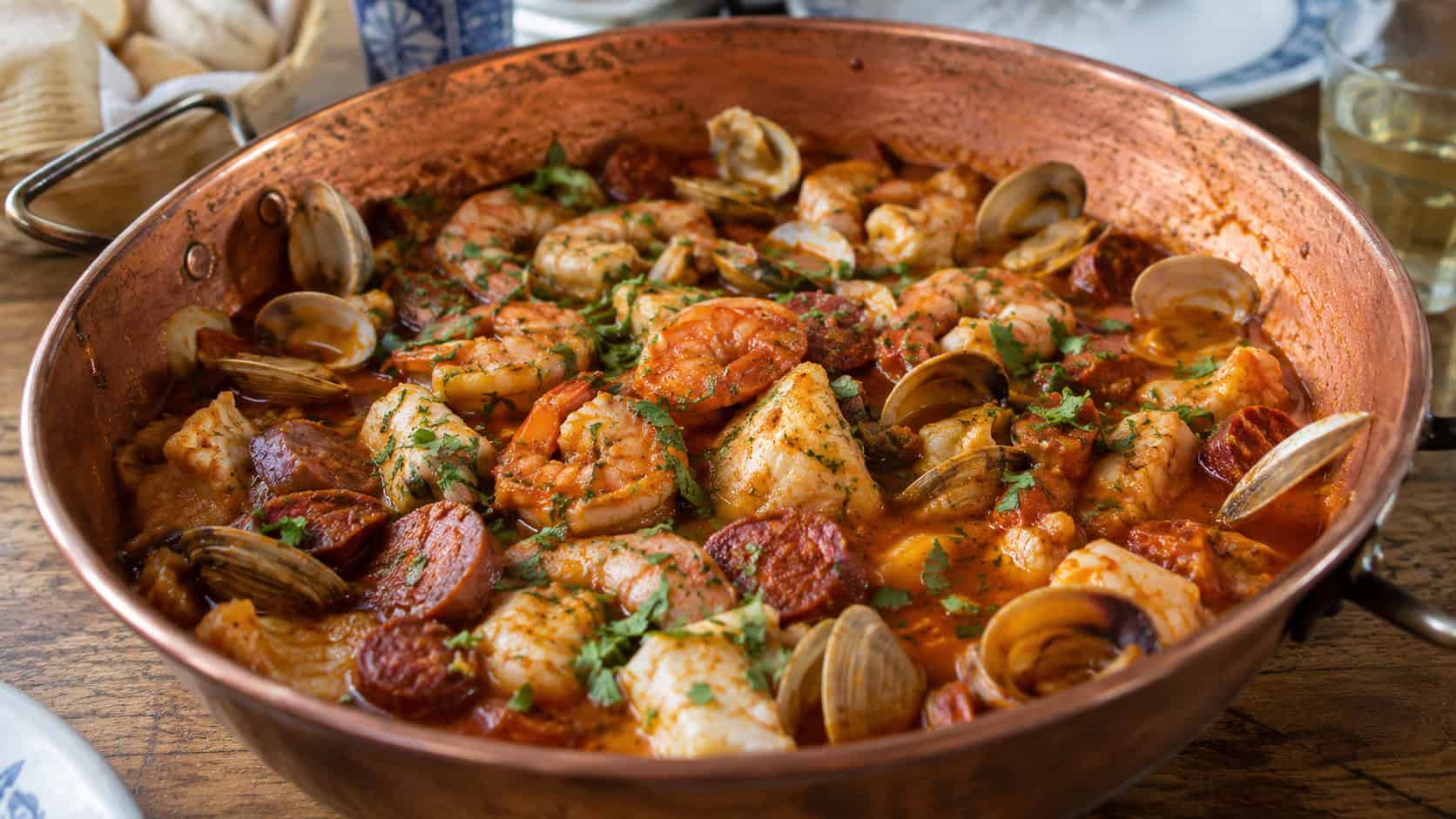 a Portuguese seafood cataplana in a wide copper pot, clams, shrimp, white fish, and chorizo in a smoky red pepper-tomato broth, garnished with cilantro, on a rustic table, natural light, shallow depth of field, landscape orientation