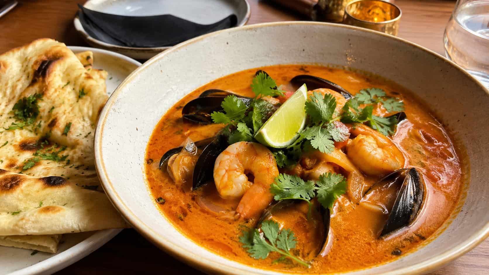 an Indian seafood curry soup in a wide bowl, shrimp and mussels in a golden coconut-tomato curry broth, topped with cilantro and lime, warm naan bread on the side, on a warm-toned table, natural light, shallow depth of field, landscape orientation