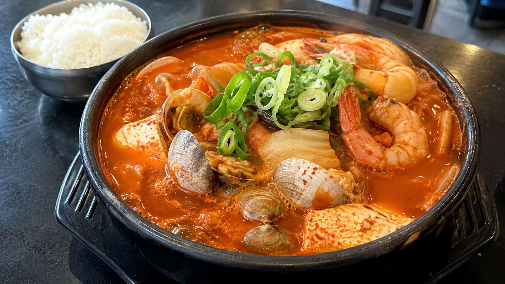 a bubbling Korean seafood jjigae in a wide stone bowl, clams, shrimp, soft tofu, and cabbage in a fiery red broth, topped with green onions, a bowl of rice nearby, on a dark table, natural light, shallow depth of field, landscape orientation