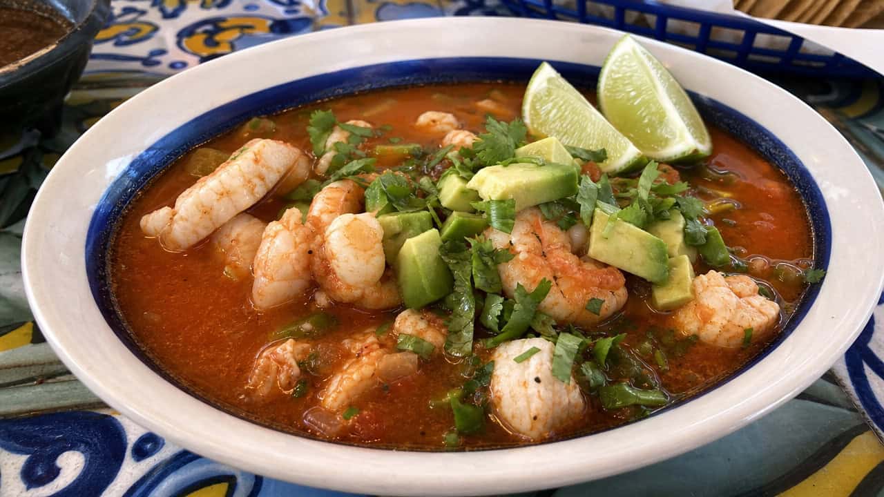 a vibrant Mexican seafood soup in a wide bowl, shrimp and white fish in a red tomato-chipotle broth, topped with cilantro, diced avocado, and lime wedges, on a colorful tiled table, natural light, shallow depth of field, landscape orientation