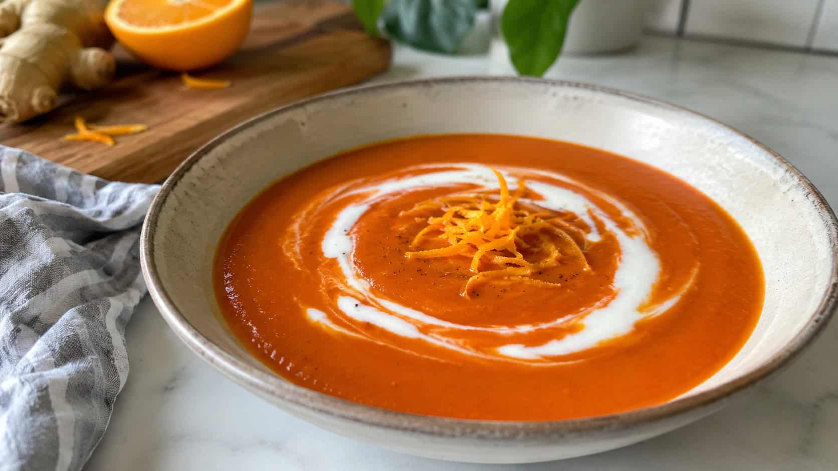 a vibrant orange-red ginger tomato soup in a wide bowl with a curl of orange zest and a swirl of coconut milk, in a wide horizontal shot, on a bright kitchen counter, natural light, shallow depth of field, landscape orientation
