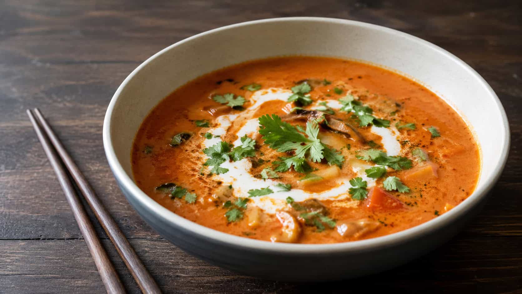 a creamy coconut Thai tomato soup in a wide bowl with a swirl of coconut milk and torn cilantro, in a wide horizontal shot, on a dark wood table with chopsticks nearby, natural light, shallow depth of field, landscape orientation